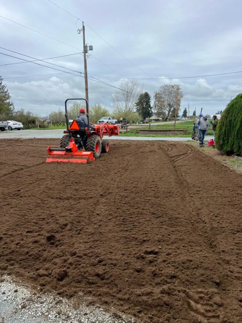 A worker operating a tractor to level soil on a large yard, preparing the area for landscaping, while other workers are seen in the background.