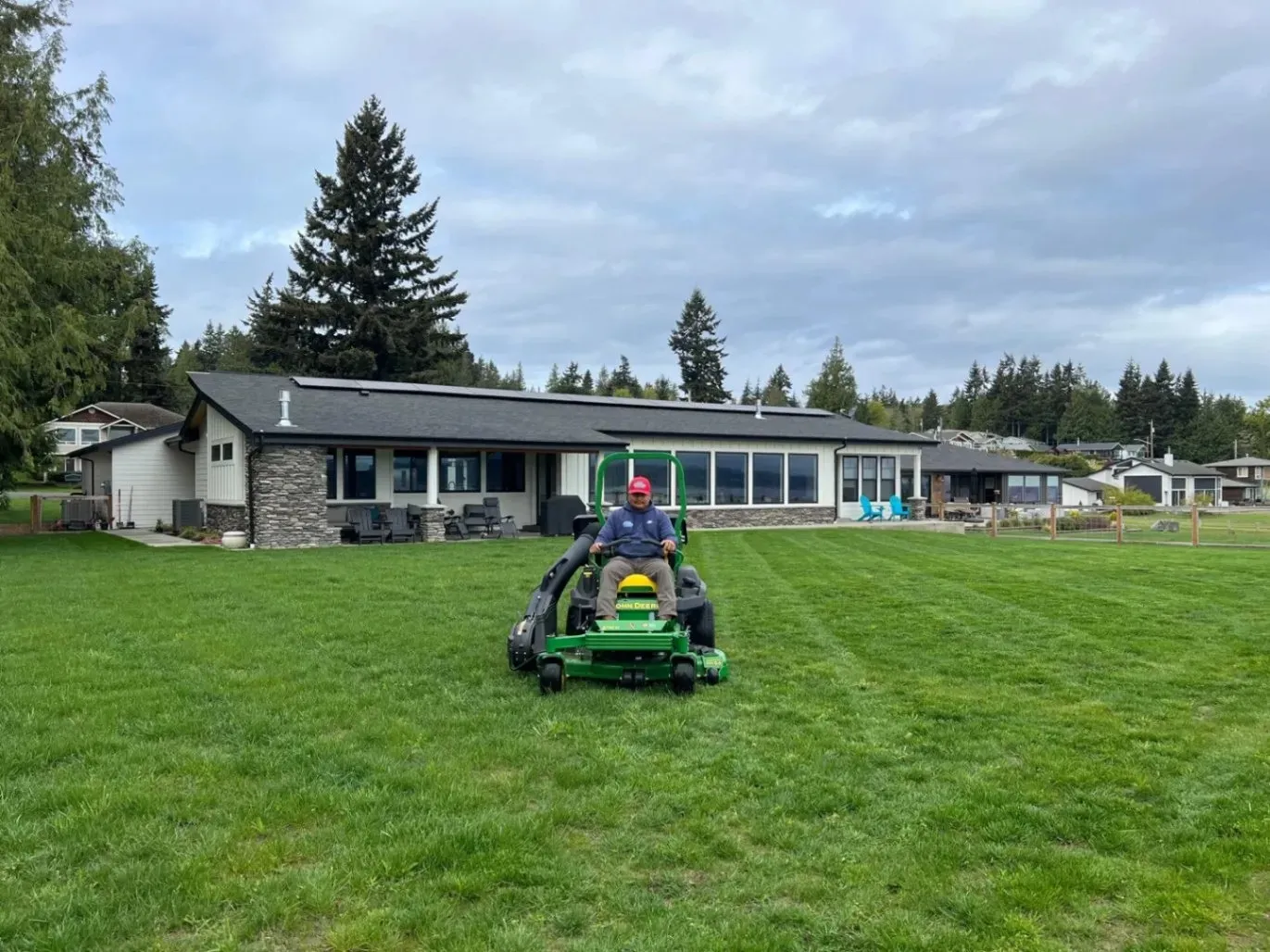 A person riding a lawn mower, cutting the grass in a large yard with a modern house and trees in the background, under a cloudy sky.