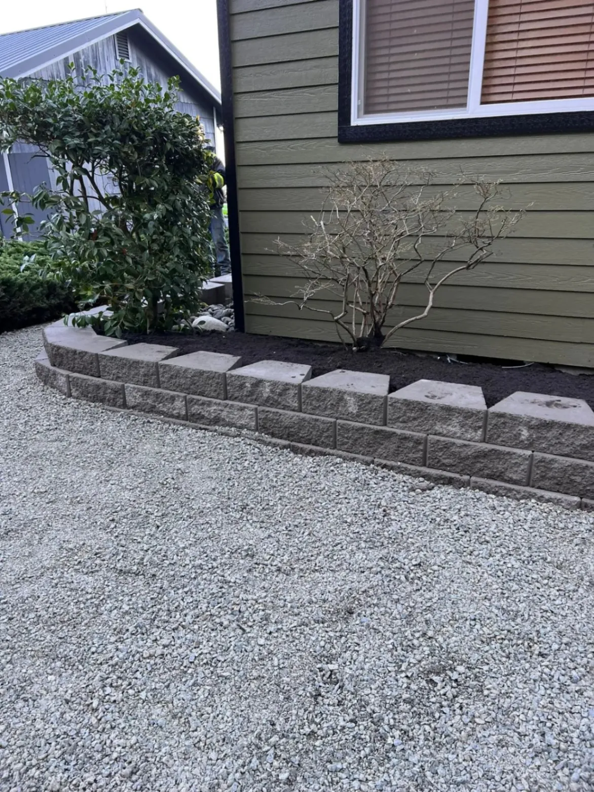 A newly built retaining wall made of concrete blocks in a landscaped yard, surrounding a small garden bed with shrubs, next to a green house with gravel on the ground.