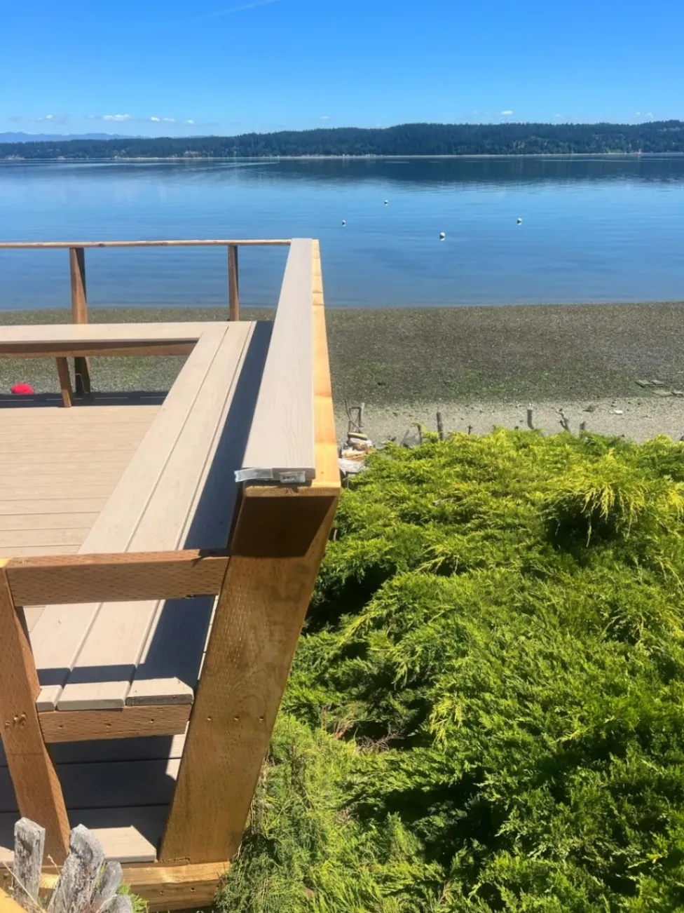 A wooden deck overlooking a calm body of water on a sunny day, with green shrubs below and distant mountains on the horizon.