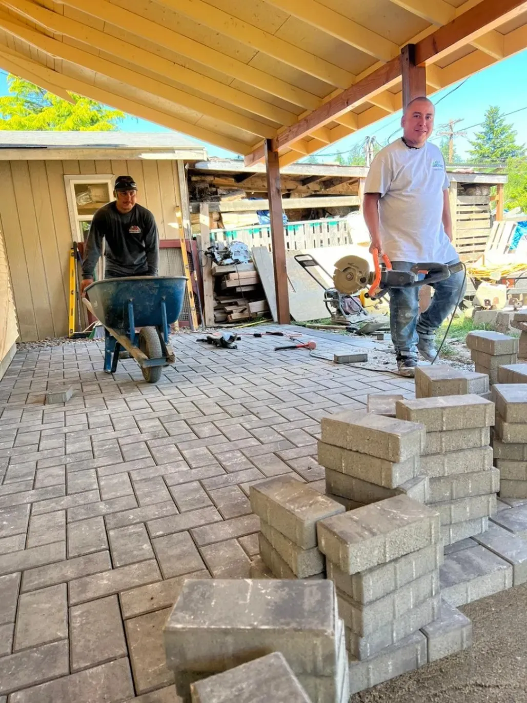 Two workers laying interlocking pavers on a patio under a wooden roof, with one worker pushing a wheelbarrow and the other holding a saw next to a stack of bricks.