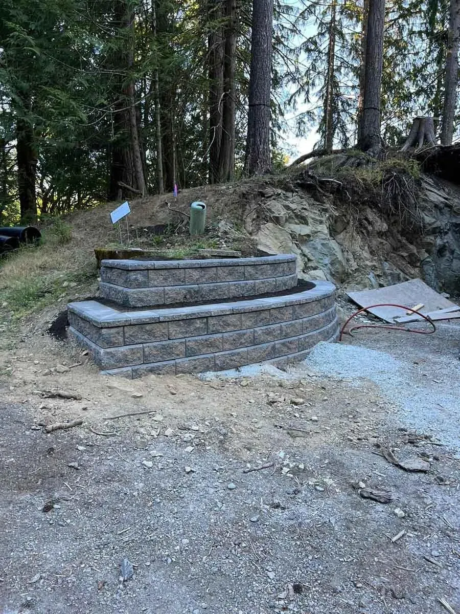 A newly constructed tiered retaining wall made of concrete blocks, built into a hillside surrounded by trees and natural terrain.