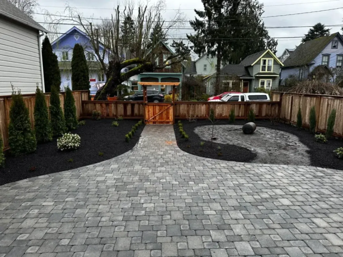 A well-maintained backyard with a paved pathway leading to a wooden gate. The landscape includes green shrubs, trees, and fresh plants surrounded by black mulch. The area has a clean and organized look with a decorative stone feature on one side of the path.