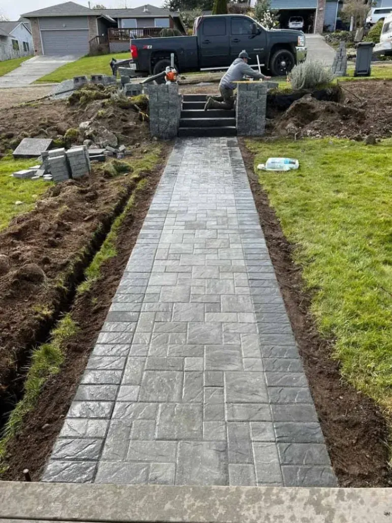 The image shows a well-constructed stone walkway with a worker laying down stones and working on the steps. The surrounding area is being prepared for further landscaping. The walkway appears to be made of textured, large stones, which adds an elegant and functional touch to the property.