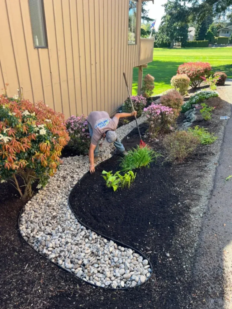 The image shows a landscaper working on a garden bed. They are arranging rocks along a curved path while planting and maintaining flowers around the edge of the garden.