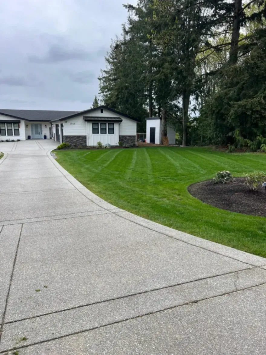 Driveway with decorative rocks, leading to a modern house with large windows, surrounded by manicured lawn and a scenic waterfront view under a cloudy sky.