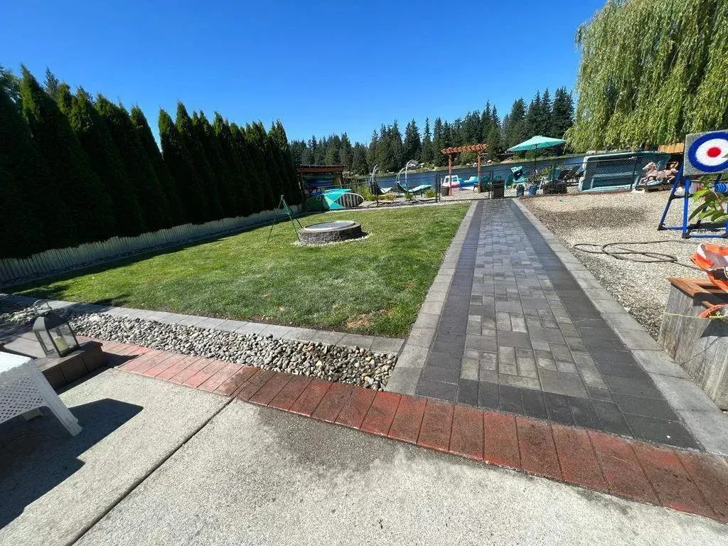 A well-maintained backyard with a paved walkway leading through a grassy area, bordered by tall evergreen trees and a decorative stone and brick path. In the background, there are outdoor seating areas, a hammock, and a playground with a swing set and a fenced pool area
