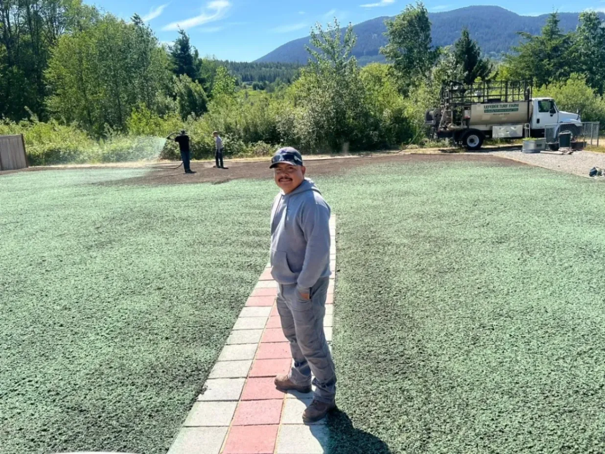 Hydroseeding services being applied to a lawn with a worker standing on a paved path, surrounded by a lush, green area. In the background, another person is operating the hydroseeding truck, ensuring an even distribution of the seed mixture.