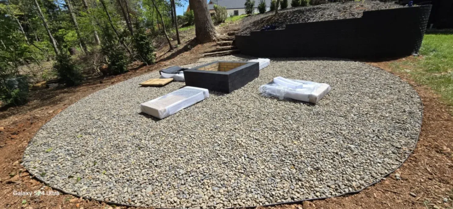 A circular gravel patio area with a square fire pit in the center and several wrapped construction materials placed around it. The space is surrounded by trees and soil, with a raised retaining wall and steps leading up a small slope in the background.