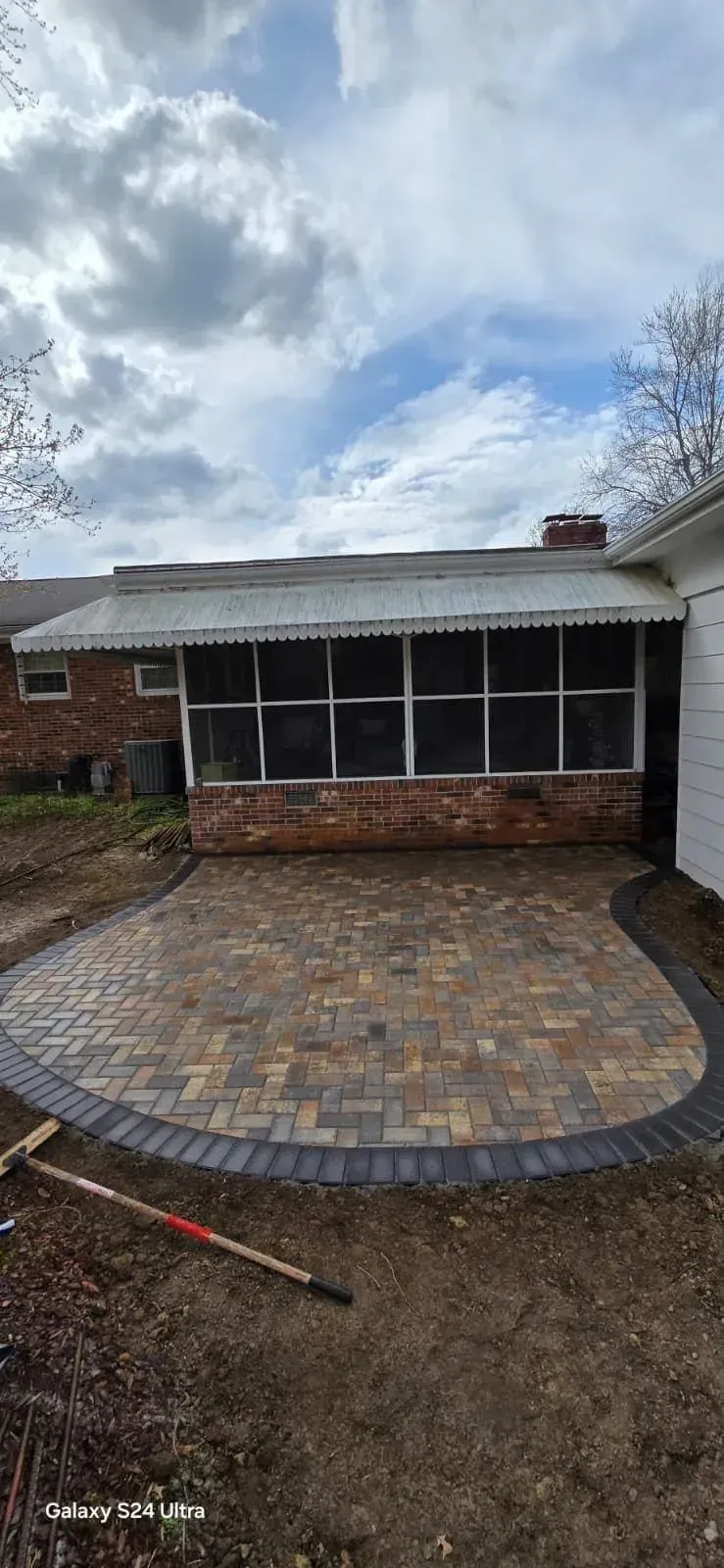 A backyard patio made of multicolored rectangular pavers with a dark border, extending from a brick house with a screened porch under a white awning. The surrounding ground is bare soil with a shovel visible in the foreground