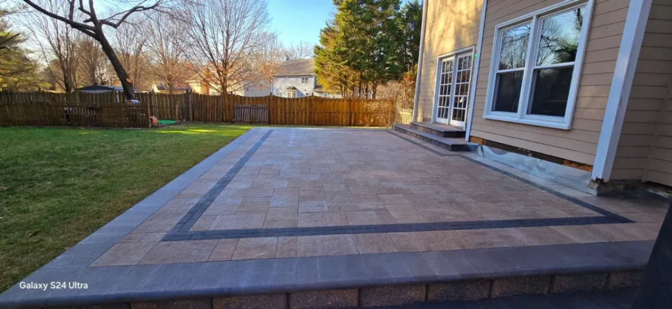 A spacious backyard patio with decorative stone pavers in a light tan color, bordered by a darker accent trim. The patio connects to a tan house with white trim and large windows, featuring steps leading to glass double doors. A wooden fence and trees line the yard in the background.
