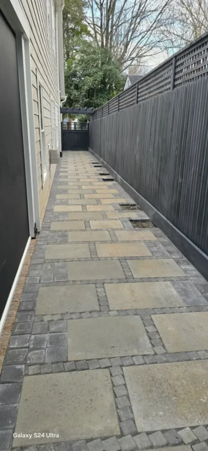 A narrow side walkway paved with large rectangular stone slabs bordered by smaller dark pavers. The path runs alongside a house with light siding and a tall black privacy fence, leading to a gate at the end. Trees and greenery are visible beyond the fence.