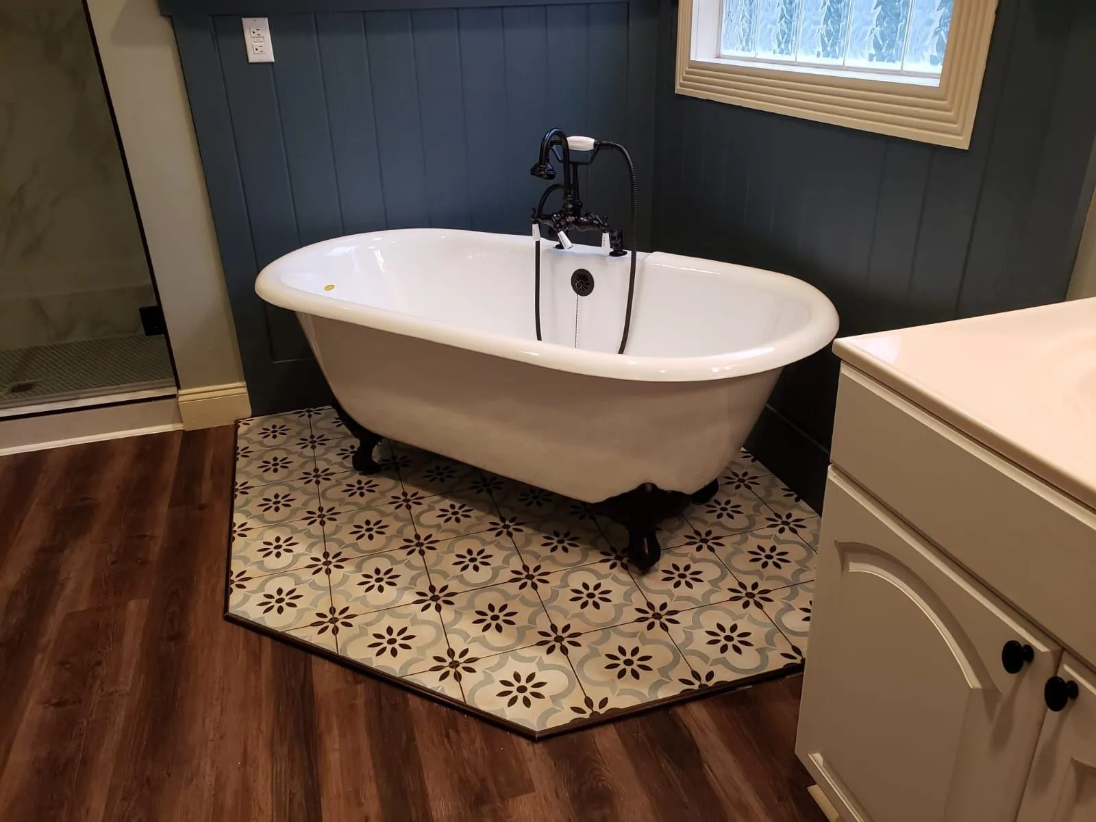 Vintage-style bathroom featuring a white clawfoot tub with black fixtures, set on a patterned hexagonal tile inlay over dark wood-look flooring. The space includes a navy shiplap accent wall, a white vanity, and a frosted window for natural light and privacy.