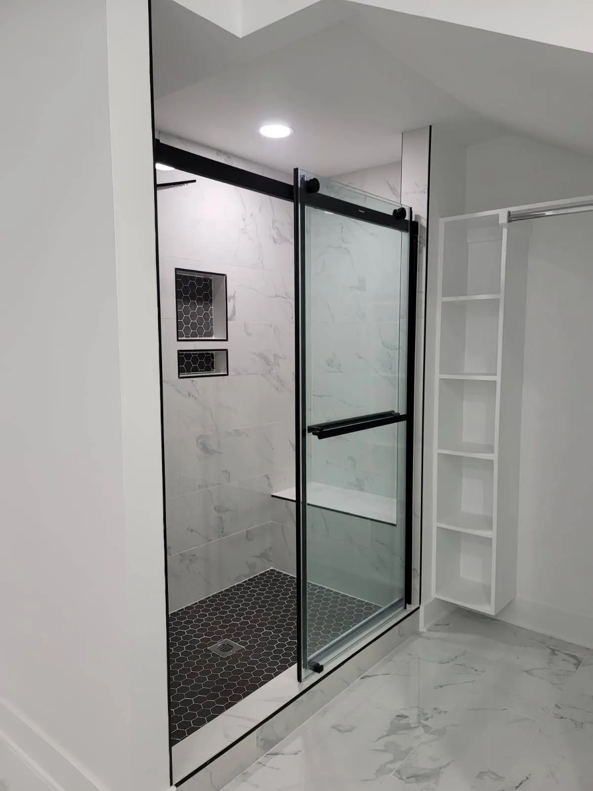 Walk-in shower installation in Raleigh, NC featuring a black hexagon tile floor, white marble-look walls, and a built-in bench. The shower includes a sliding glass door with black trim and dual recessed niches with dark hex tile backing for added contrast and storage.