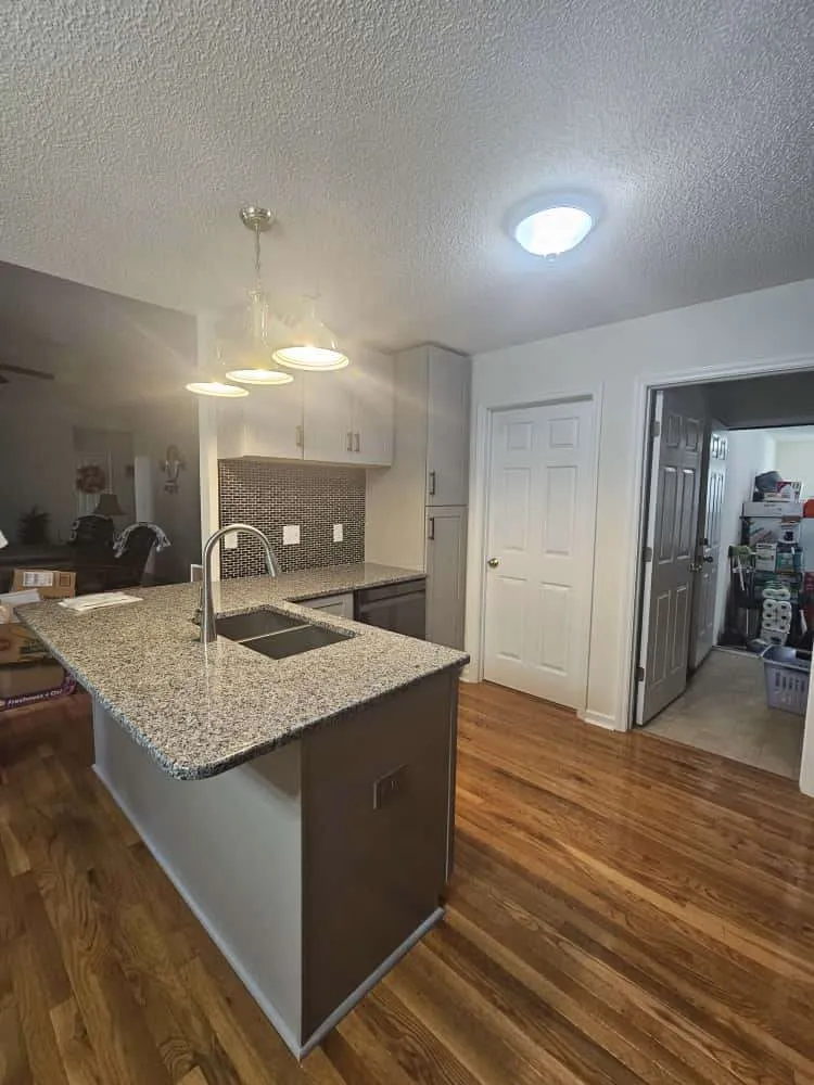 Modern kitchen with polished hardwood kitchen flooring, granite countertops, and a gray island with built-in sink and seating. The space features a mosaic tile backsplash, pendant lighting, and an open layout connected to adjacent rooms.