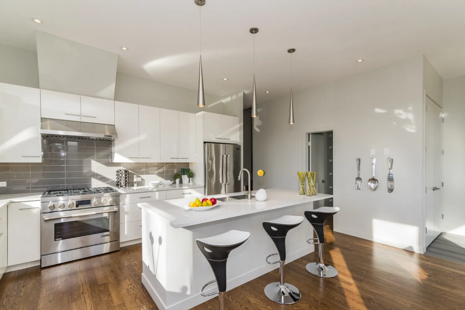 Bright kitchen with open-concept design featuring sleek white cabinets, stainless steel appliances, and a central island with a modern waterfall countertop. Pendant lighting and barstools complete the airy layout with warm wood flooring throughout.