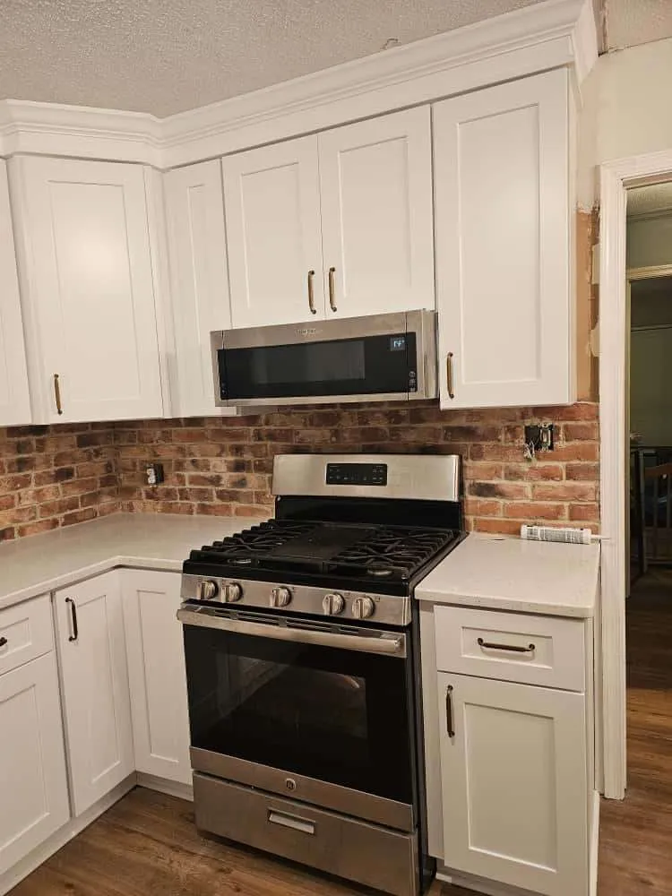 Custom kitchen design featuring white shaker cabinets, quartz countertops, and a warm exposed brick backsplash. A stainless steel gas range and built-in microwave are centered in the layout, creating a functional and stylish cooking area.