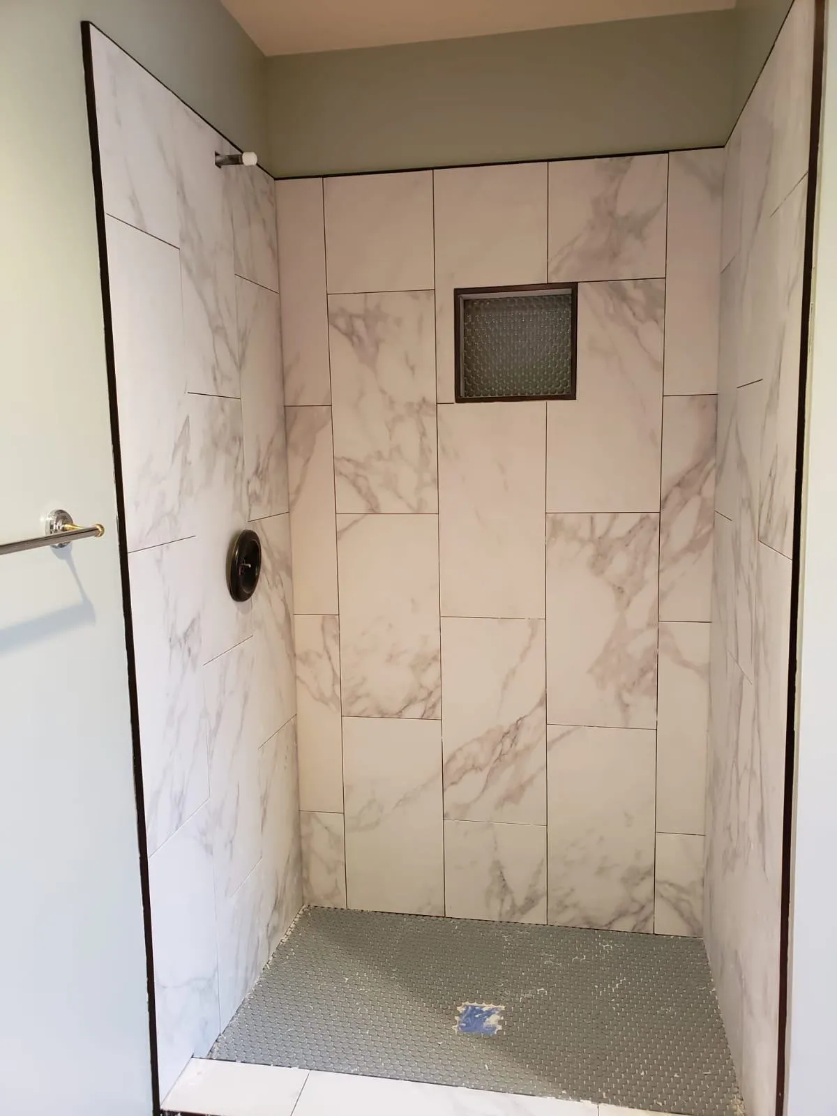 Tile shower installation in progress featuring large rectangular white tiles with gray marble veining and a black-edged recessed niche. The unfinished floor is prepped with a blue drain and small mosaic tile base, ready for final installation.