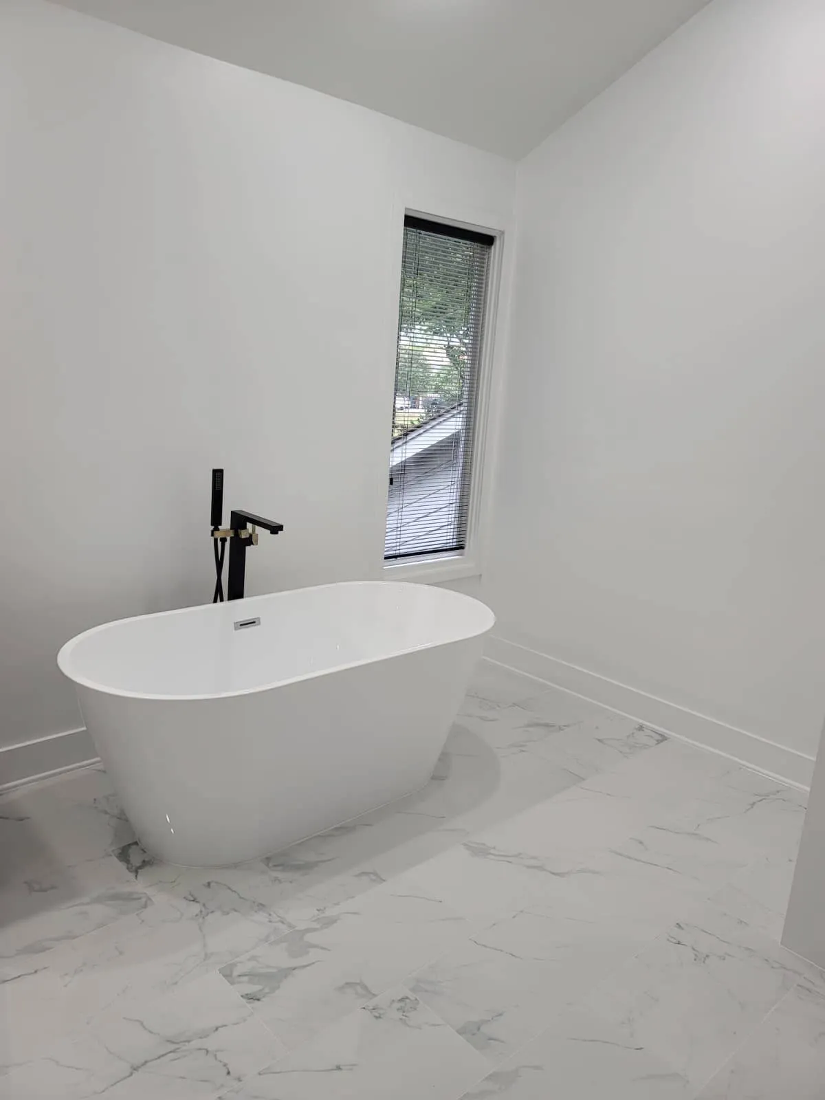 Sleek bathroom renovation showcasing a modern freestanding white tub with a matte black floor-mounted faucet. The space features glossy marble-look tile flooring and a tall, narrow window bringing in natural light against clean white walls.