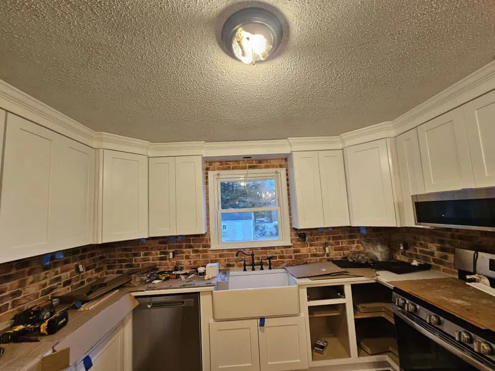 Kitchen remodel in progress by skilled kitchen designers in Northwest Raleigh, featuring white shaker cabinets, exposed brick backsplash, and a farmhouse sink beneath a central window. Construction tools and protective tape indicate active installation of appliances and countertops.