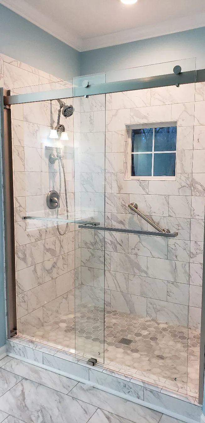 Tub-to-shower conversion featuring a frameless sliding glass door, white marble-style tiles with gray veining, and a small built-in window. The walk-in shower includes a handheld showerhead, corner shelf, and stainless steel grab bar for added safety and accessibility.