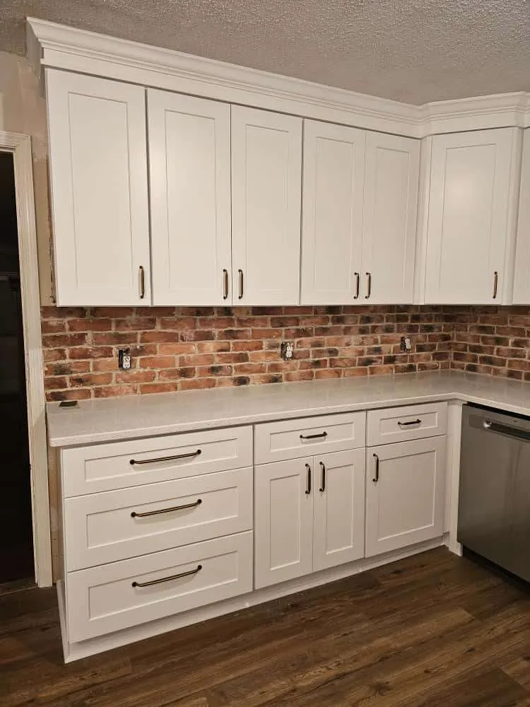 Custom cabinet installation featuring white shaker-style upper and lower cabinets with dark hardware, paired with a warm exposed brick backsplash. Light quartz countertops and wood-look flooring complete the transitional kitchen design.