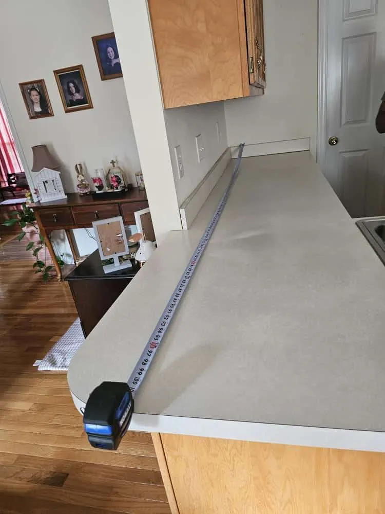 Home kitchen with a laminate countertop being measured for replacement, indicating plans to upgrade your kitchen with premium countertops. A tape measure runs the length of the surface, capturing dimensions in a cozy, traditionally styled interior.