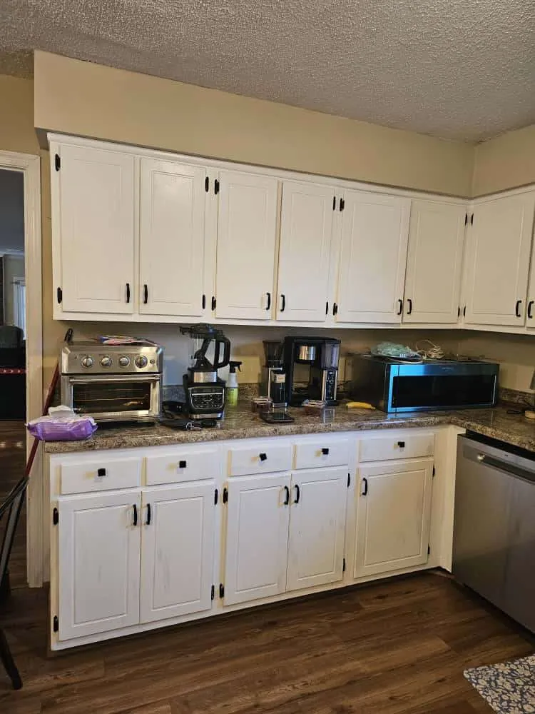 Older kitchen with white custom cabinet installations featuring black hardware and granite countertops. Small appliances like a coffee maker, microwave, and toaster oven line the counter, highlighting a functional but well-used space.