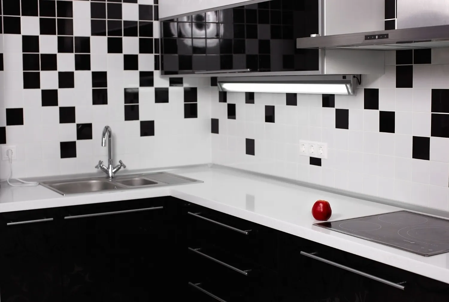 Modern kitchen with a custom kitchen backsplash featuring a bold black and white checkerboard tile pattern. Sleek black cabinetry, a minimalist cooktop, and a single red apple on the white countertop create a sharp, contemporary aesthetic.