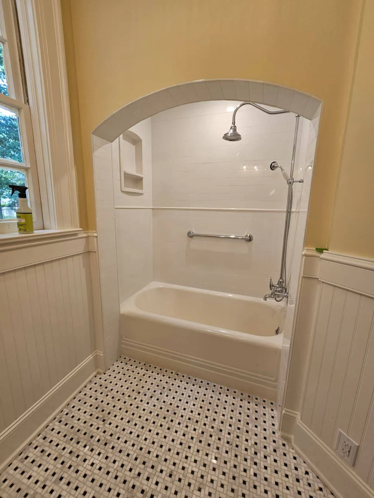 Traditional-style bathroom renovation in Raleigh, NC featuring a white alcove bathtub with vintage-style fixtures and an arched tiled shower surround. The space includes a built-in shower niche, grab bar for safety, and black-and-white mosaic tile flooring for a classic finish.