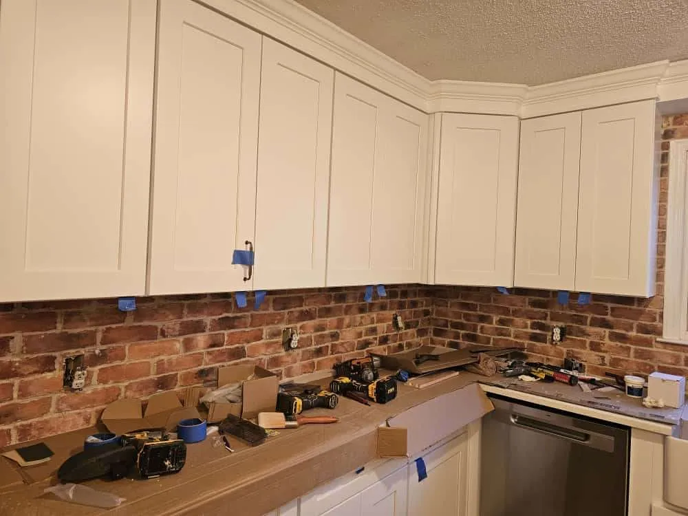 In-progress kitchen remodel focused on maximizing your kitchen’s potential with smart storage solutions, featuring newly installed white shaker cabinets and a brick backsplash. Tools and painter’s tape line the counters, indicating ongoing work to enhance storage and functionality.