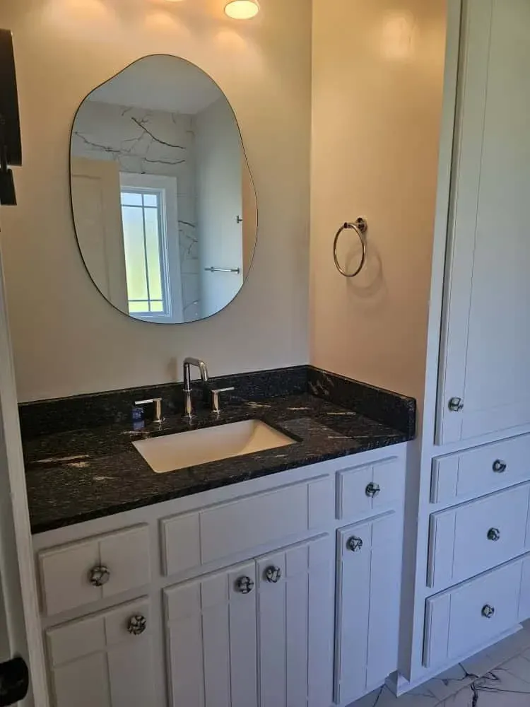 Bathroom upgrade showcasing a white vanity with a black granite countertop, under-mount sink, and polished chrome faucet. The space features a uniquely shaped mirror, updated recessed lighting, and built-in cabinetry offering enhanced storage solutions.