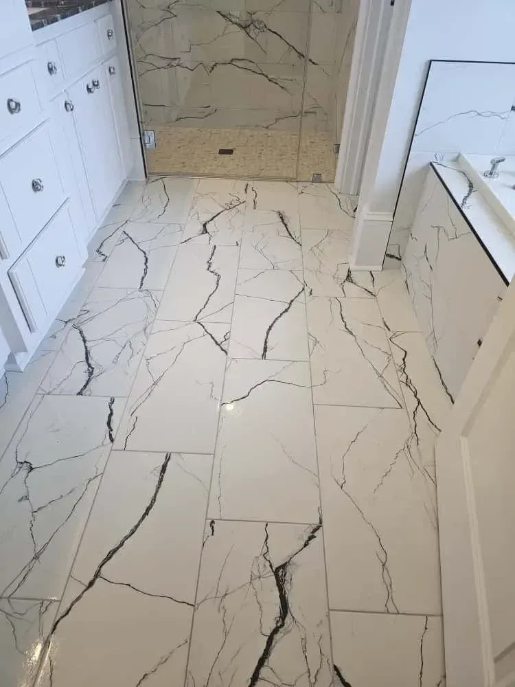 Bathroom flooring installation featuring large white tiles with dramatic black veining in a marble-look pattern. The modern design extends throughout the space, complementing the glass-enclosed shower and white cabinetry with chrome hardware.