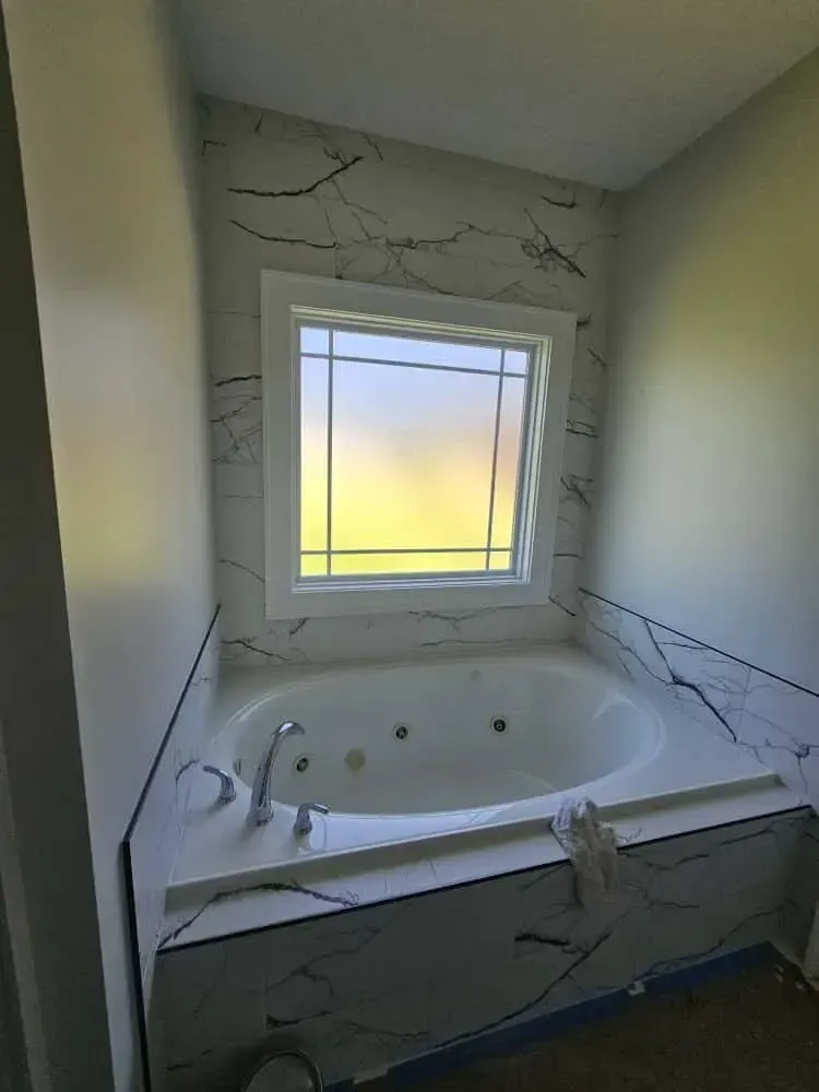 Bathroom remodel featuring a built-in jetted tub with chrome fixtures, surrounded by white marble-look tile with dramatic black veining. A frosted window above the tub allows natural light while maintaining privacy.