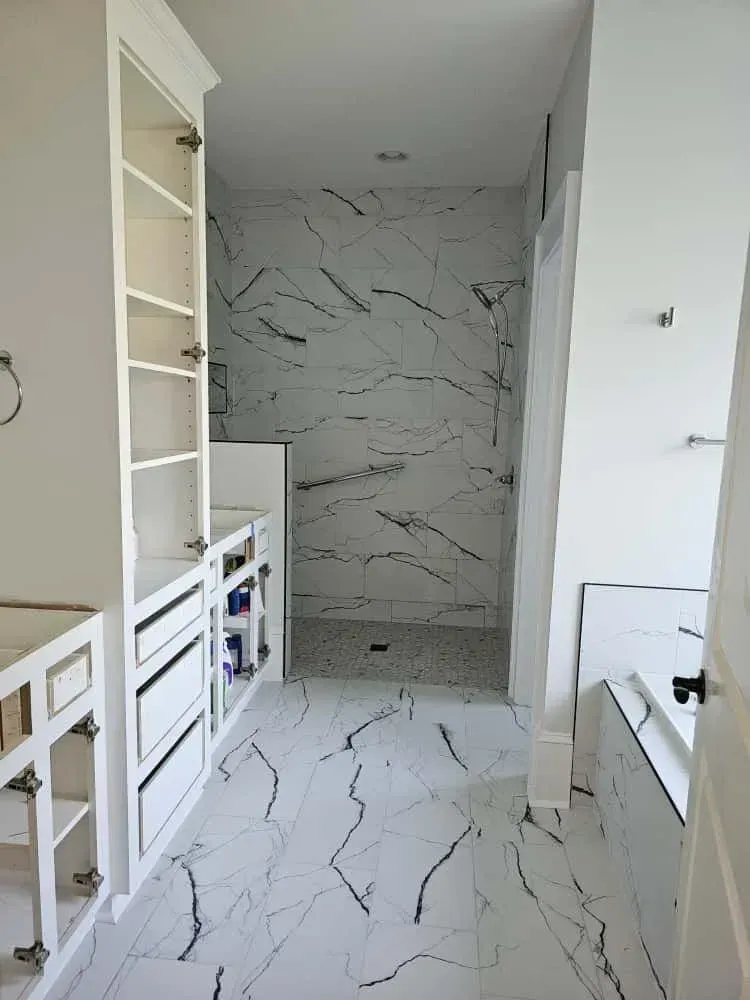 Bathroom renovation in progress featuring bold marble-look floor tiles, open white cabinetry, and a walk-in shower with matching large-format wall tiles. The shower floor is finished with small mosaic tiles, and a soaking tub with black trim accents is built into the corner.