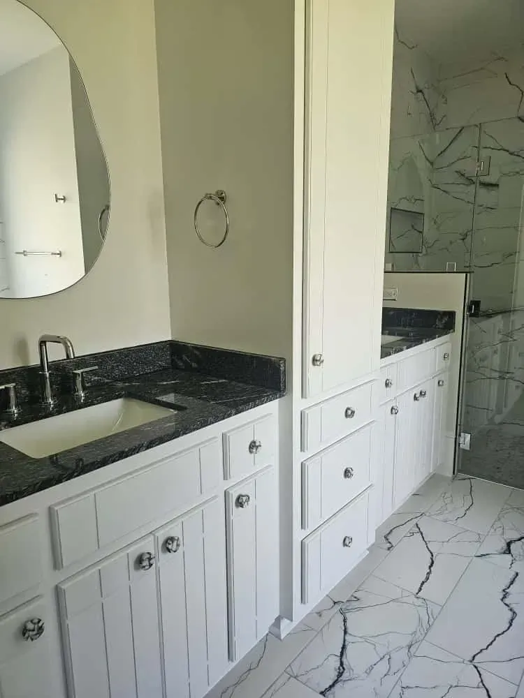 Luxury fixture installation featuring dual vanities with white cabinetry, black granite countertops, and polished chrome faucets. The bathroom includes marble-look tile flooring with bold veining and a glass-enclosed shower with matching wall tiles for a cohesive, upscale design.
