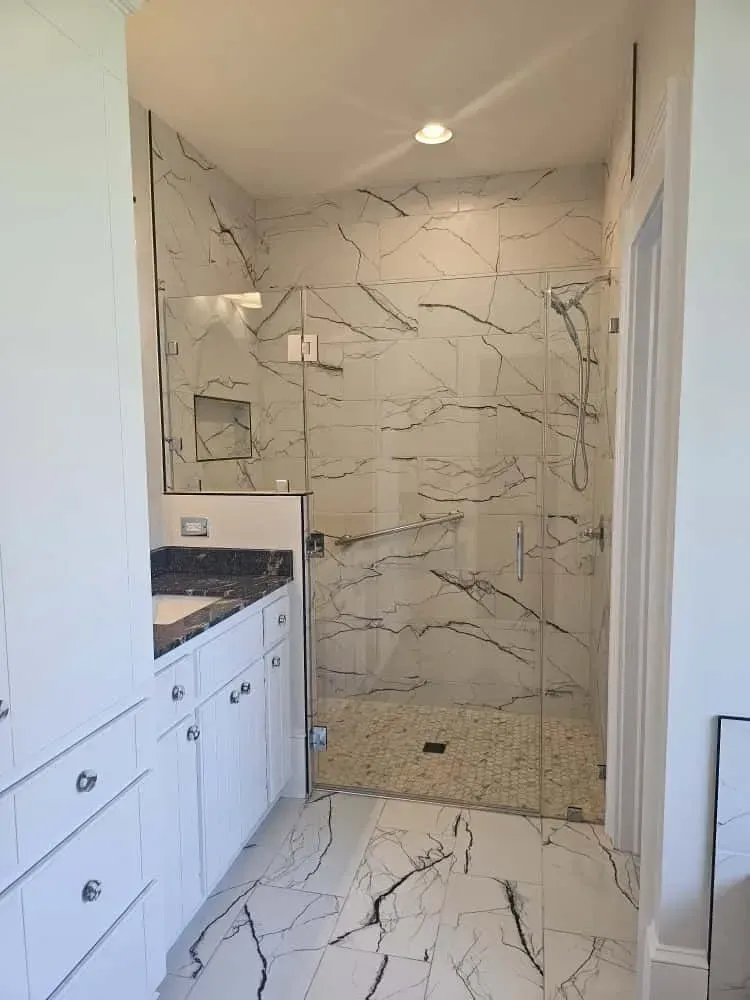 Luxurious bathroom featuring a frameless glass walk-in shower with large marble-look tiles, a handheld showerhead, and a safety grab bar. The space includes a white vanity with a dark granite countertop, chrome hardware, and matching marble-look flooring for a cohesive design.