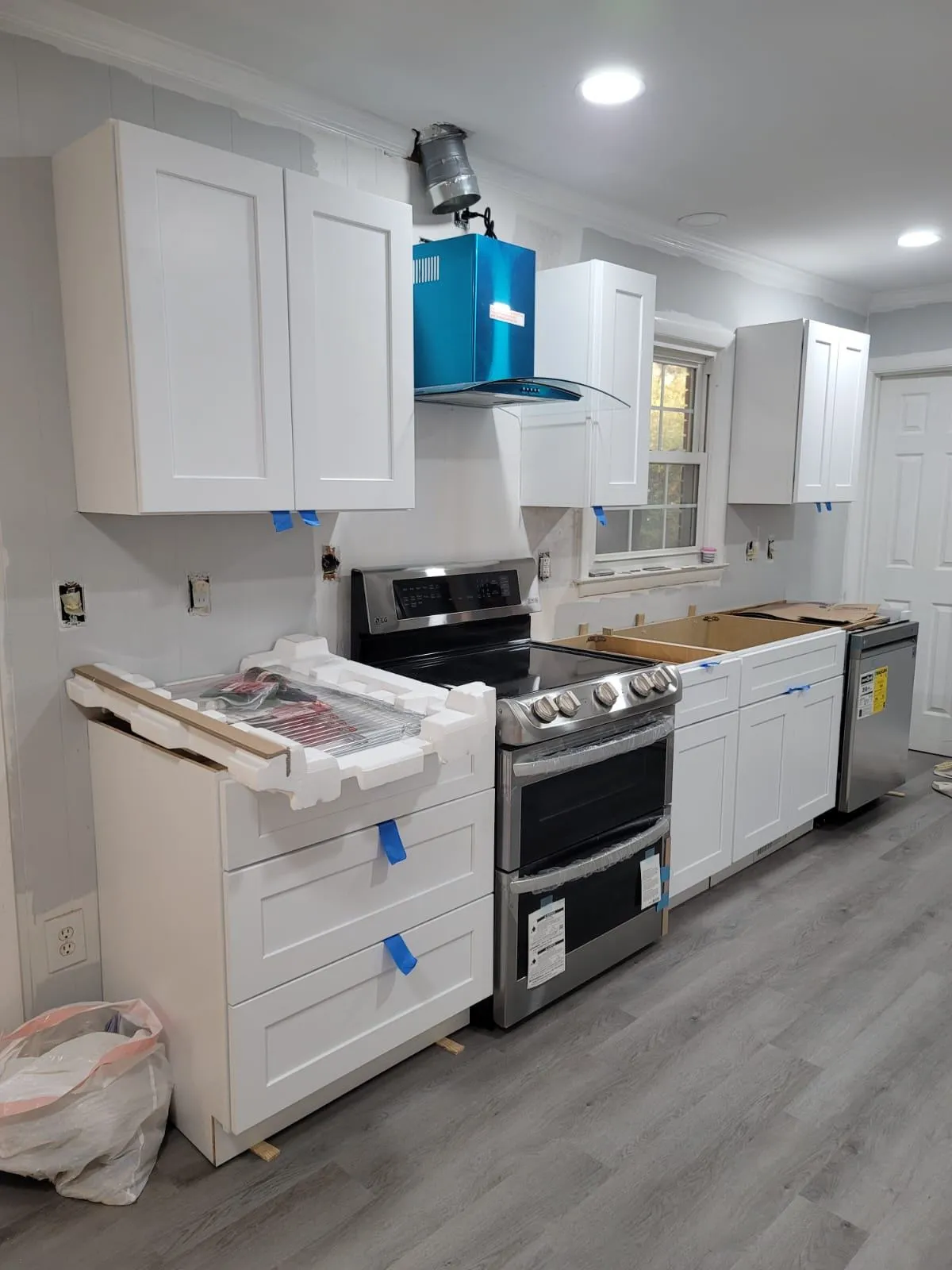 In-progress affordable kitchen renovation in North Raleigh featuring newly installed white shaker cabinets, a stainless steel stove and dishwasher, and protective wrap on drawers and appliances. Gray plank flooring and under-cabinet lighting add a modern touch to the partially finished space.