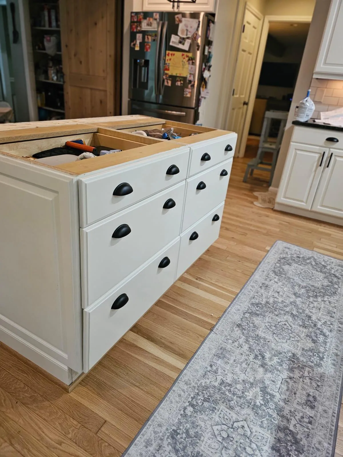 In-progress kitchen island installation showcasing custom cabinetry with white shaker-style drawers and matte black cup pulls. The drawer boxes are exposed, filled with tools, sitting atop light wood flooring beside a patterned gray runner.