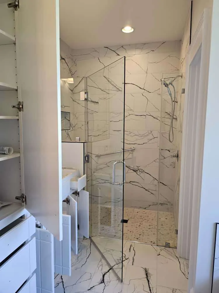 Modern full bathroom renovation featuring a frameless glass shower with white marble-style tile and bold black veining. Open cabinet doors and drawers reveal white storage units, adding functional space to the sleek, clean design.