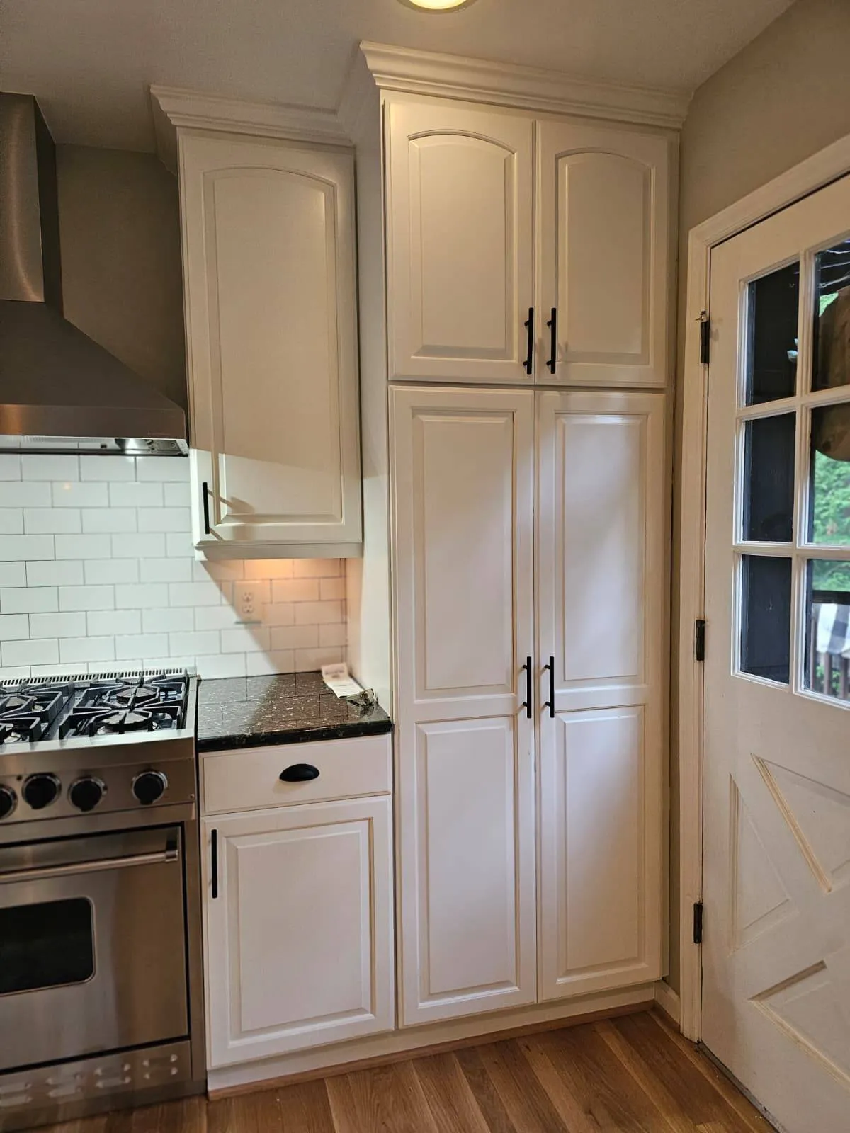 Custom cabinet installation featuring white shaker-style upper and lower cabinets with dark hardware, paired with a warm exposed brick backsplash. Light quartz countertops and wood-look flooring complete the transitional kitchen design.