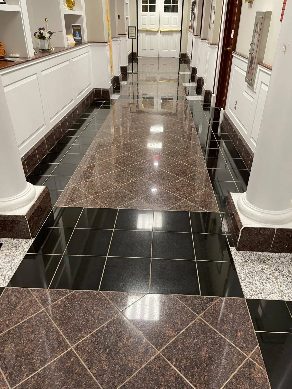 A polished hallway with glossy black and brown tiled floors, white walls with decorative paneling, and columns along the sides, leading to double doors at the end.