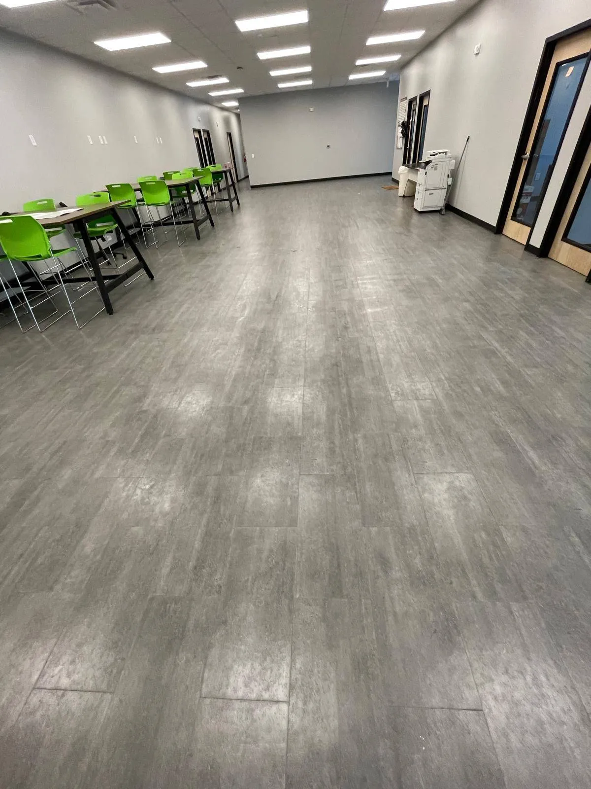 Spacious office breakroom with gray flooring, green chairs, tables, and bright overhead lighting