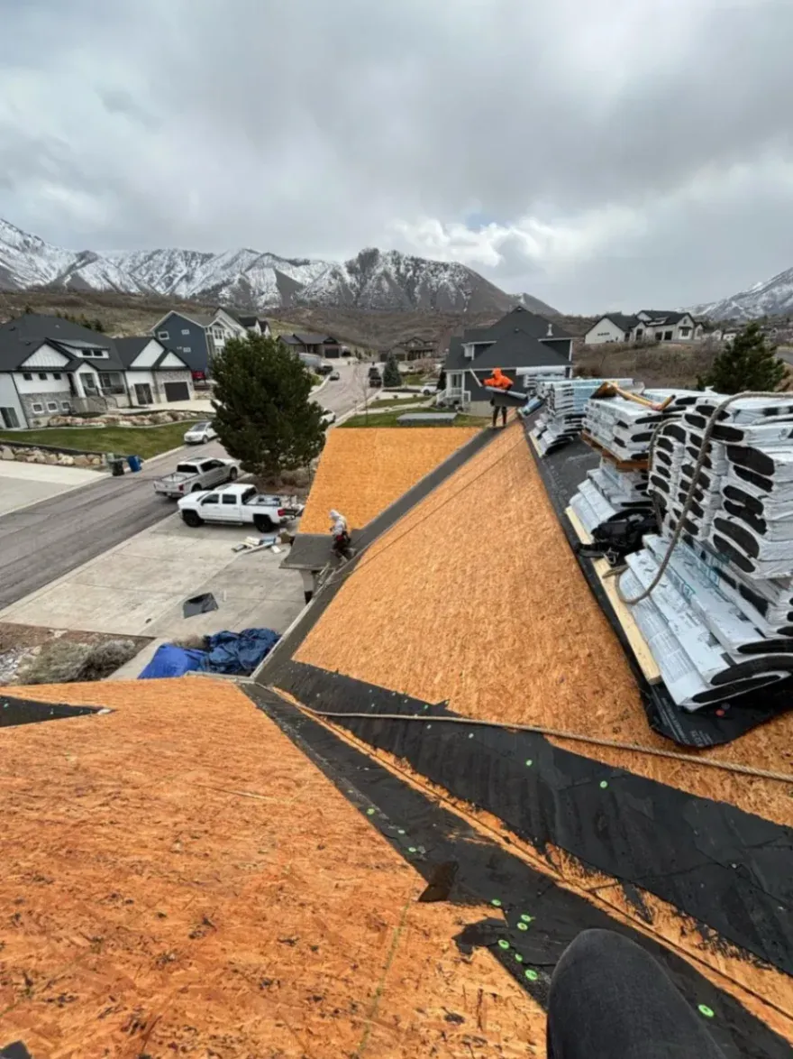 Close-up of a roof under construction, featuring newly installed plywood sheathing and roofing felt, with workers preparing for shingle installation, set against a mountainous backdrop.