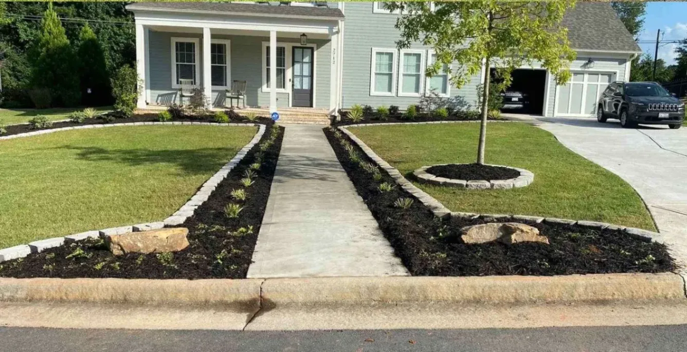 A neatly landscaped front yard in Milton, GA, with a straight concrete walkway, fresh mulch beds with young plants, stone edging, and a circular tree bed enhancing curb appeal.