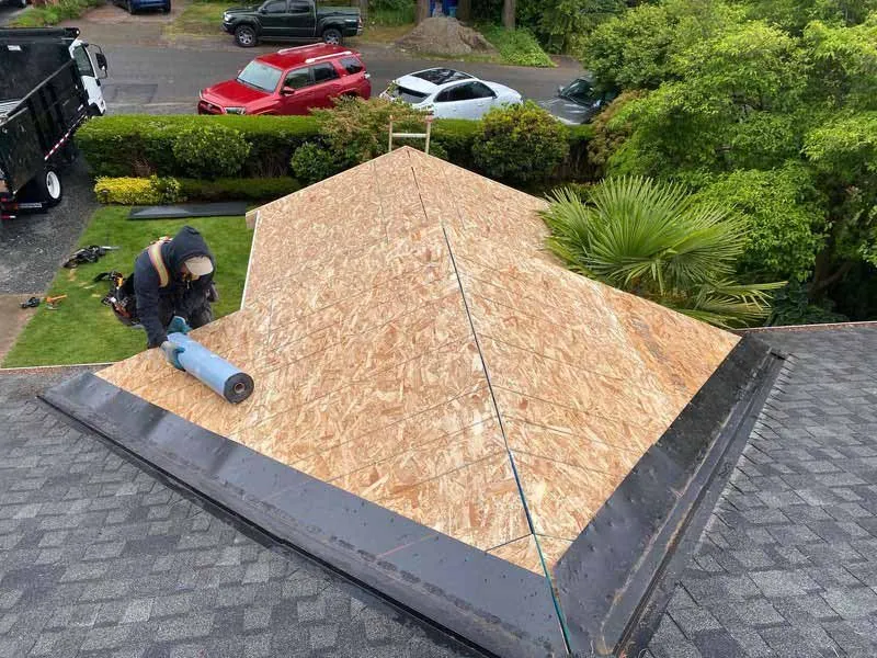 Man installing roofing material on a partially completed roof with plywood and shingles, surrounded by greenery