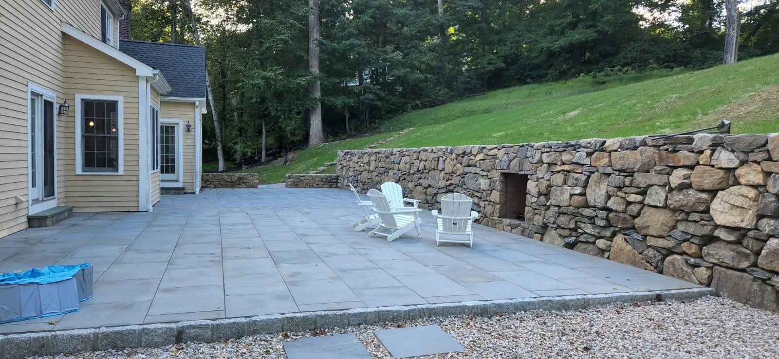 A spacious stone patio with large square pavers and white Adirondack chairs, bordered by a natural stone retaining wall, located in New Canaan, CT.