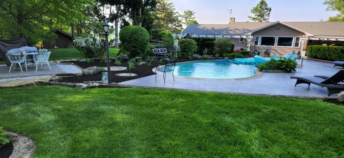 A landscaped backyard with a curved in ground pool, manicured lawn, and neatly trimmed shrubs surrounding a patio seating area. Lawn Care Services near Westport, CT