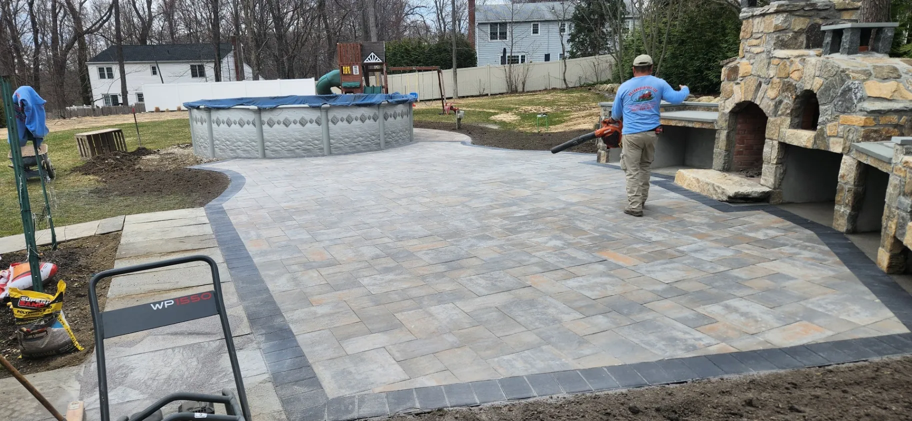 A newly installed paver patio with a dark border beside a stone outdoor fireplace and above ground pool, completed by our landscape designers in New Milford, CT.