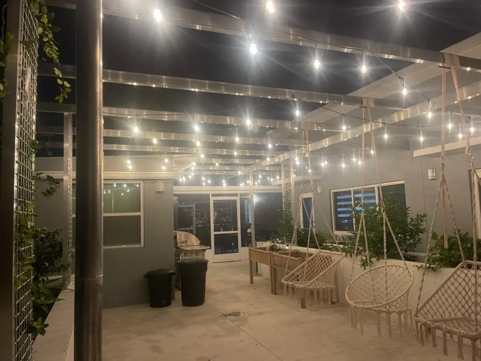 A custom metal trellis patio structure fabricated by Abel Welding Services in Davie, FL, illuminated by overhead string lights and featuring suspended macrame swing chairs.